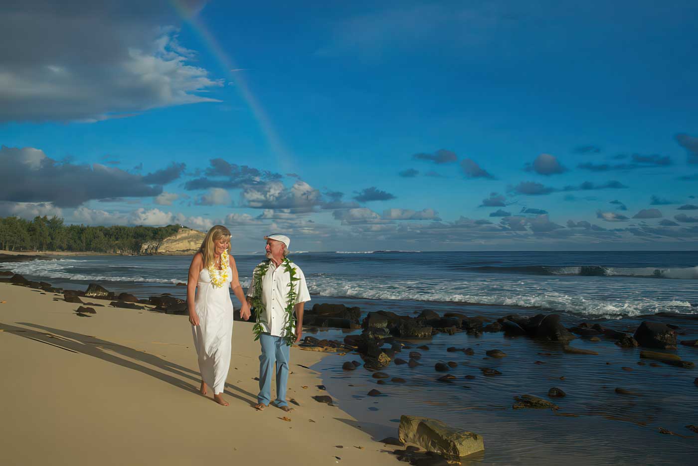 Couple walking hand-in-hand along a sandy beach under a rainbow, dressed in white and adorned with Hawaiian lei. Kauai South Shore Weddings.