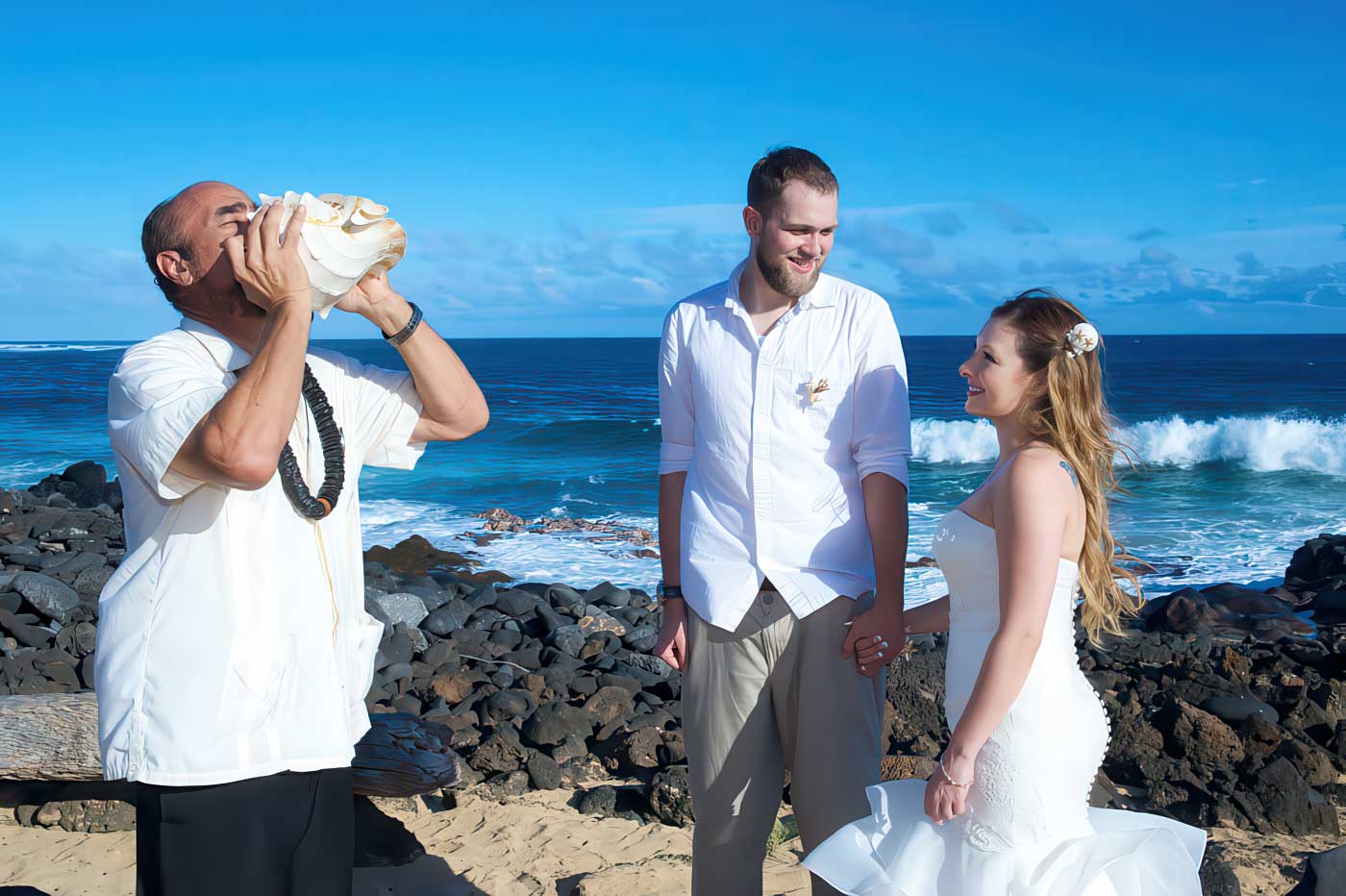 Pastor Kilborn blowing a conch shell as bride and groom stand together by the ocean during their Hawaiian beach wedding ceremony. Kauai South Shore Weddings.