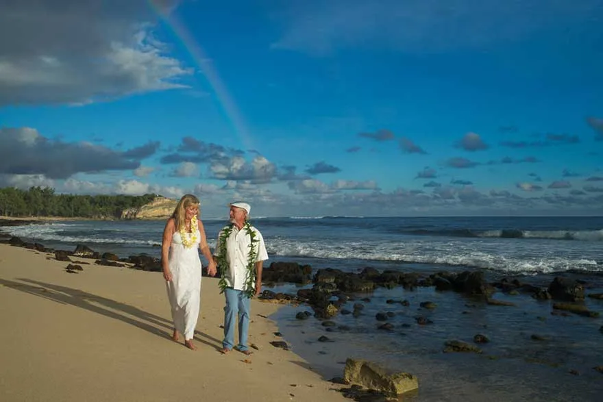 Smiling couple in wedding attire walking barefoot along a rocky beach on Kauai's South Shore, with a rainbow arching across a bright blue sky. Kauai South Shore Weddings with Pastor Kilborn.