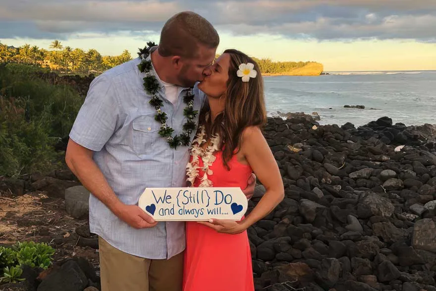 
A couple kisses by the ocean at sunset, holding a sign that says "We (Still) Do and always will...". A beachside vow renewal performed by Pastor Kilborn of Kauai South Shore Weddings.